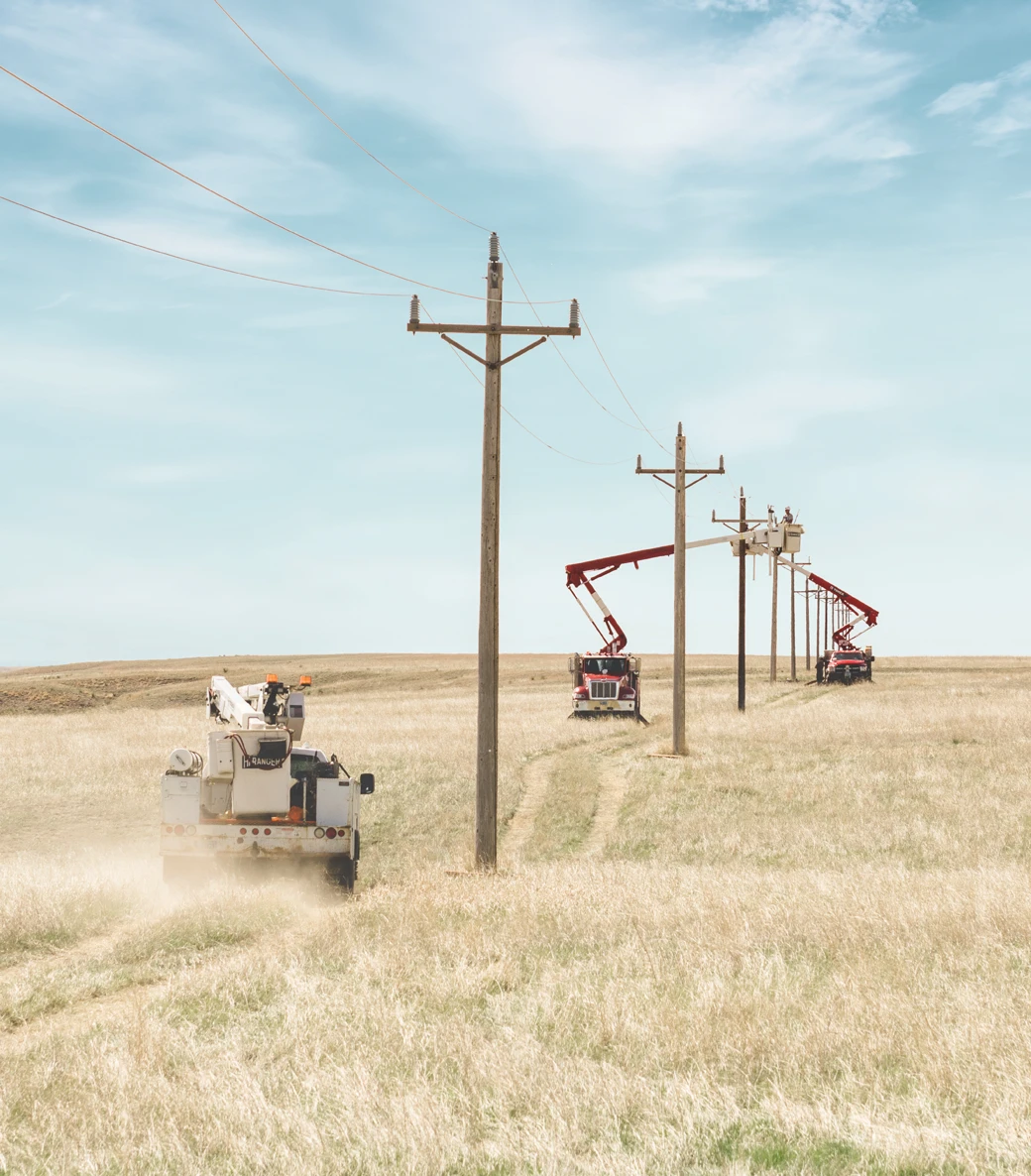 Utility trucks with elevated work platforms are parked along a rural field, with workers servicing or inspecting power lines on wooden utility poles under a clear sky.