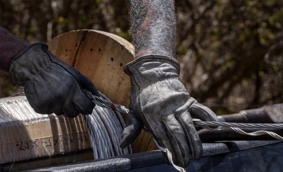 Two gloved hands handle thick metal cables near a large wooden spool outdoors.