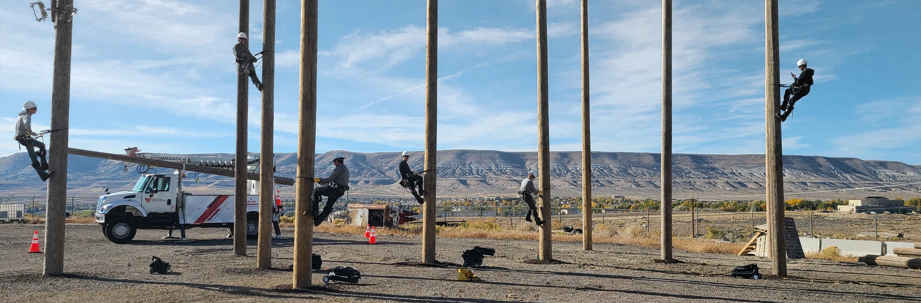 Six utility workers in safety gear climb tall wooden poles in an outdoor training area, with a utility truck and mountainous landscape in the background.