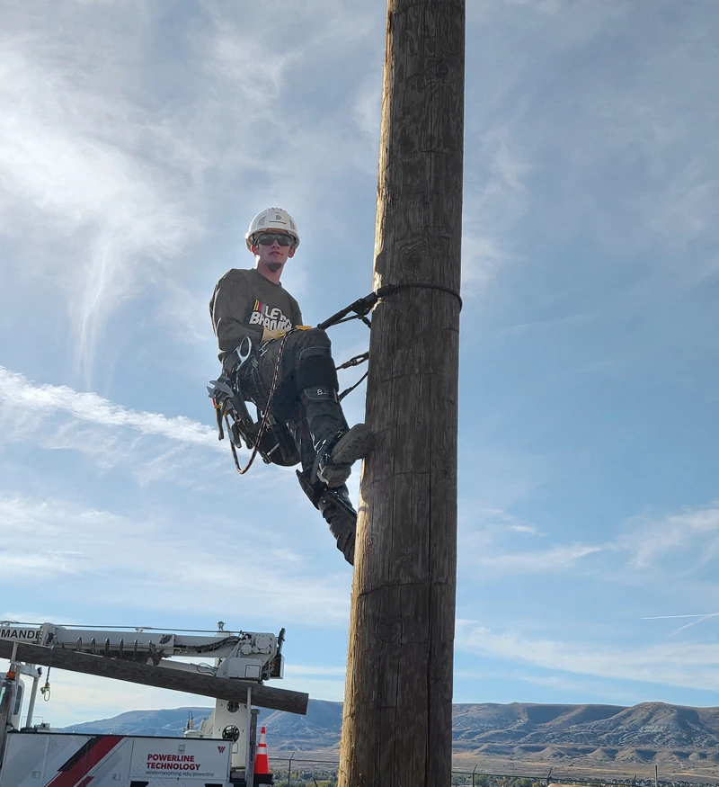 A utility worker in safety gear is climbing a wooden pole using a harness, with a service truck and mountains in the background.