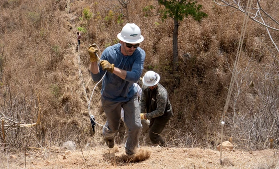 Two workers wearing helmets and gloves climb a steep, dry hill using ropes, surrounded by brown grass and sparse vegetation.