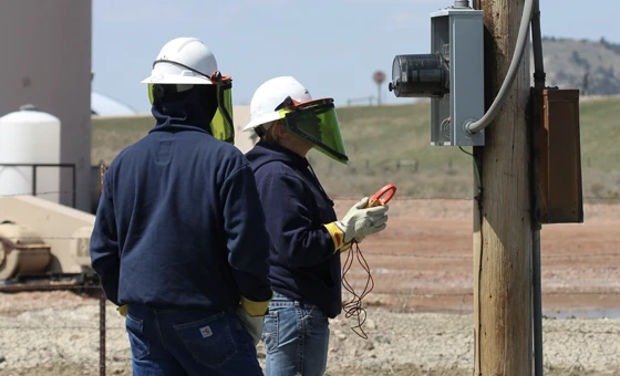Two workers wearing protective gear stand outdoors by an electrical panel; one uses a handheld meter to check the equipment.