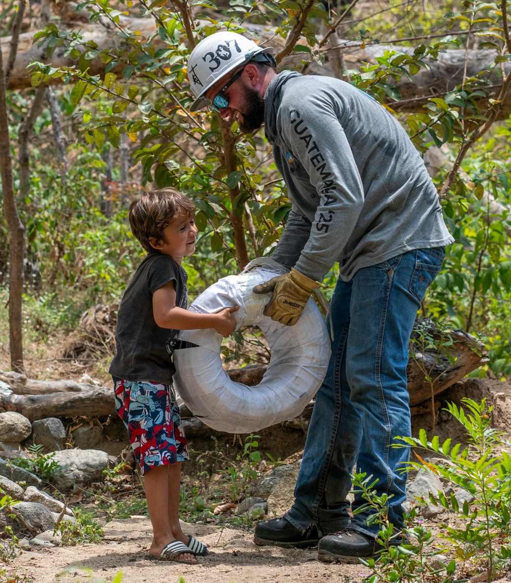 A man in work gear helps a young boy hold a large spool of cable outdoors in a wooded area.