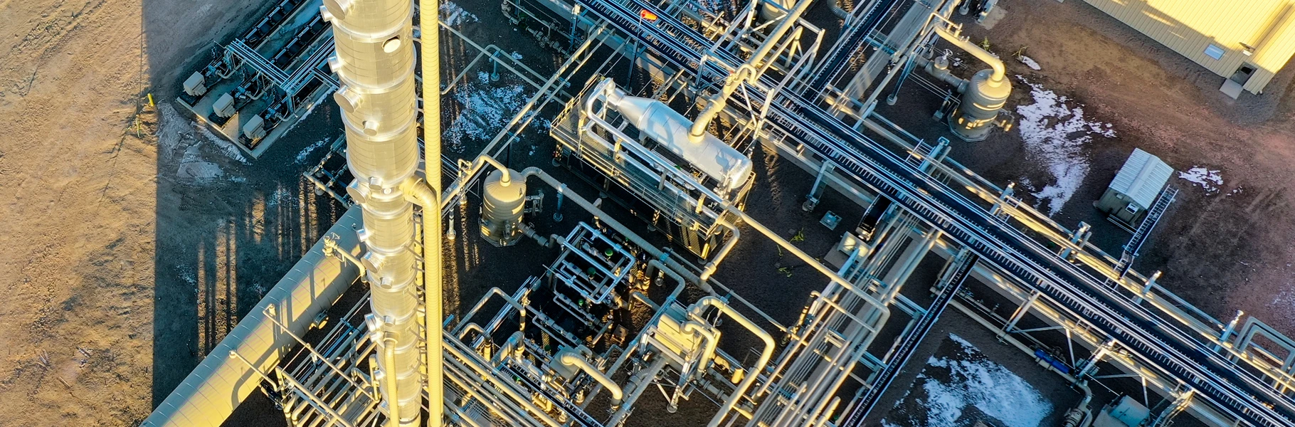 Aerial view of an industrial facility with numerous pipes, tanks, and structures, some snow patches on the ground, and part of a building visible in the top right corner.