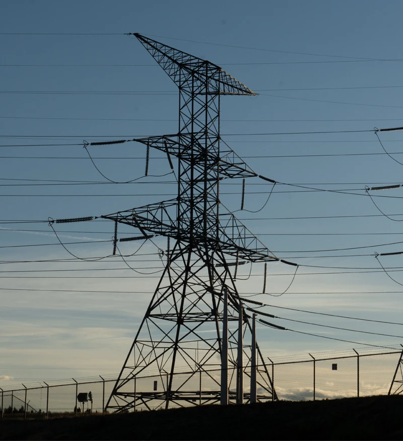 A high-voltage electricity pylon with multiple power lines is seen against a blue sky, with a fence at the base in the foreground.