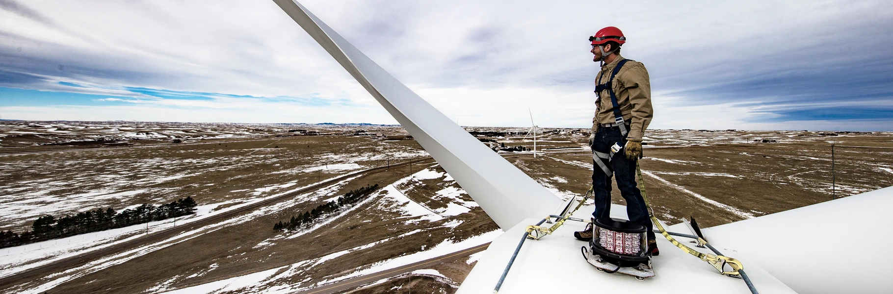 A worker wearing safety gear and a red helmet stands on top of a wind turbine blade, overlooking a vast, snow-dusted landscape with open fields and cloudy skies.