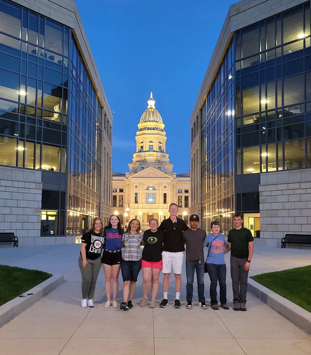 Eight people stand in a row outside between two modern buildings, with a lit domed capitol building in the background at dusk.